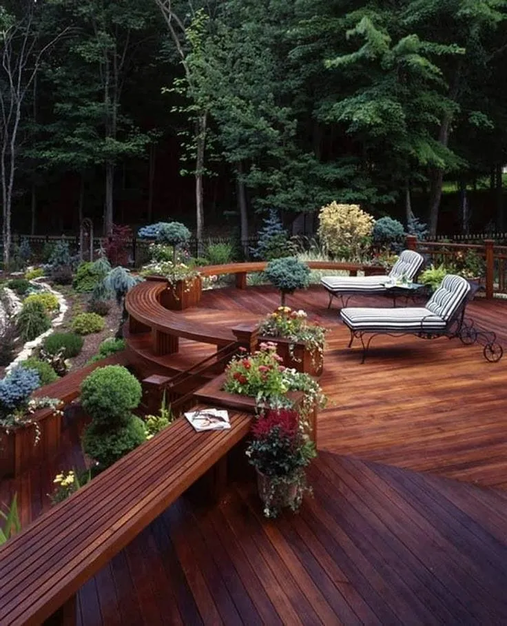 This curved deck surrounded by mature trees and layered planting shows how powerful natural landscaping can be. Instead of trying to tame the space, the deck design follows the shape of the garden, wrapping gently around shrubs, planters, and existing greenery. The built-in seating and planter boxes blur the line between deck and garden, making the whole area feel tucked away and private without needing screens or fencing. This approach works beautifully for wooded backyards or larger plots where you want the deck to feel like a retreat rather than a feature dropped into the space. It’s a great reminder that sometimes the best deck landscaping idea is simply working with what’s already there.