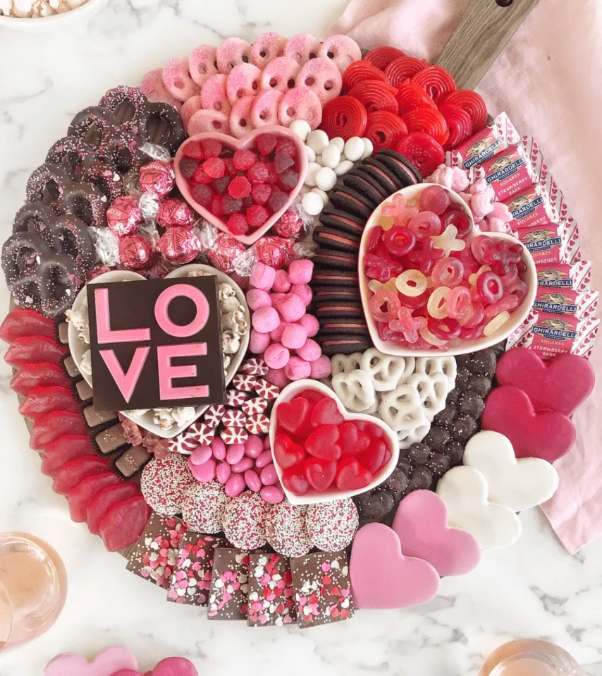 This pink food board is a candy lover’s dream, styled in soft blush, red, and white tones for a fun Valentine’s vibe. It’s packed edge-to-edge with chocolate-covered pretzels, strawberry wafer cookies, gummy hearts, marshmallows, sandwich cookies, white chocolate treats, and wrapped chocolate squares. Heart-shaped bowls filled with gummies and chocolates anchor the center, while layered sweets arranged in sections give the board that full, abundant look without feeling messy. It’s playful, colorful, and perfect for a party spread where everyone can grab their favorites.